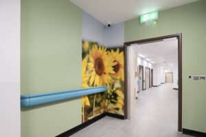 A hospital corridor, the closest walls are light green with a large print of some sunflowers. The distant part of the corridor is white with a number of doors leading off.