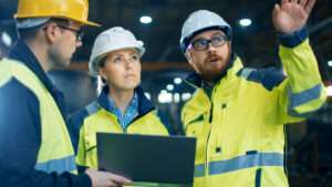 Three people in high visibility jackets and hard hats, having a conversation. One of them is holding a small laptop computer.