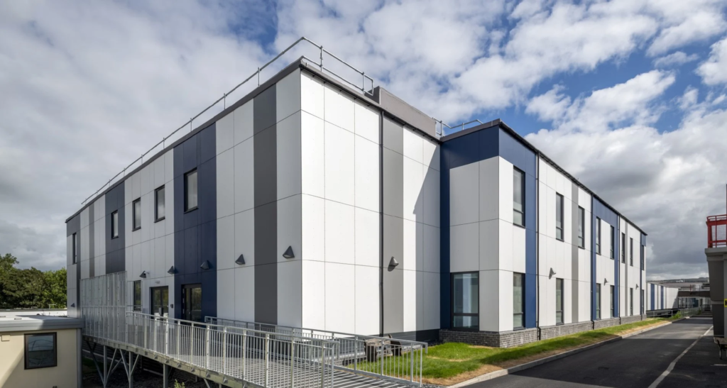 External three-quarter view of the two-storey hospital building at Airedale. The external classing is white with blue and grey stripes and there are windows at regular intervals on both floors. To the left is the building entrance, with a long, gentle ramp running up to the doors. In the foreground and leading off to the right is a tarmac walkway. The sky is blue and dotted with white clouds.