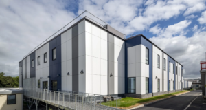 External three-quarter view of the two-storey hospital building at Airedale. The external classing is white with blue and grey stripes and there are windows at regular intervals on both floors. To the left is the building entrance, with a long, gentle ramp running up to the doors. In the foreground and leading off to the right is a tarmac walkway. The sky is blue and dotted with white clouds.