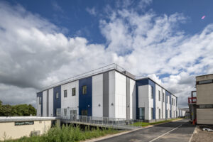 External three-quarter view of the two-storey hospital building at Airedale. The external classing is white with blue and grey stripes and there are windows at regular intervals on both floors. To the left is the building entrance, with a long, gentle ramp running up to the doors. In the foreground and leading off to the right is a tarmac walkway. The sky is blue and dotted with white clouds.