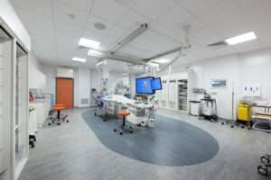 A wide angle view of a cardiac catheterisation laboratory/ At the rear there is an orange door. Storage units and medical equipment are around three sides of the room. In the centre there is a bed for patients along with a range of diagnostic equipment.