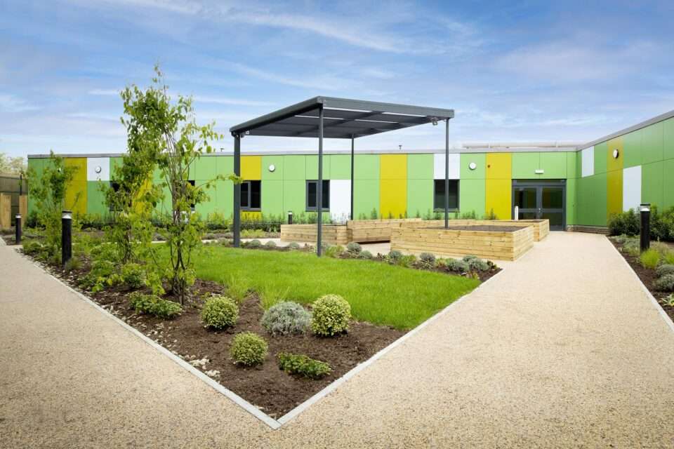 A garden area featuring raised beds, low level borders, and a pergola. In the background is the ward building, with green, yellow and white cladding panels. The sky is blue and mostly clear.