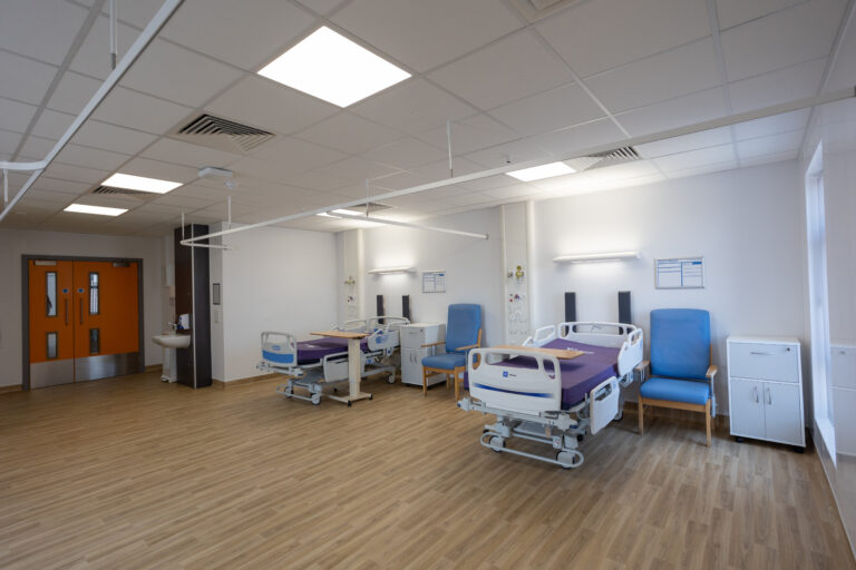 Inside a ward room. To the left is an orange double door. To the centre and right or the image are two bays with a hospital bed, tall backed chair and bedside cabinet in each bay. There is a window to the far right. The walls and ceiling are white, the floor is a beech colour wood effect vinyl.