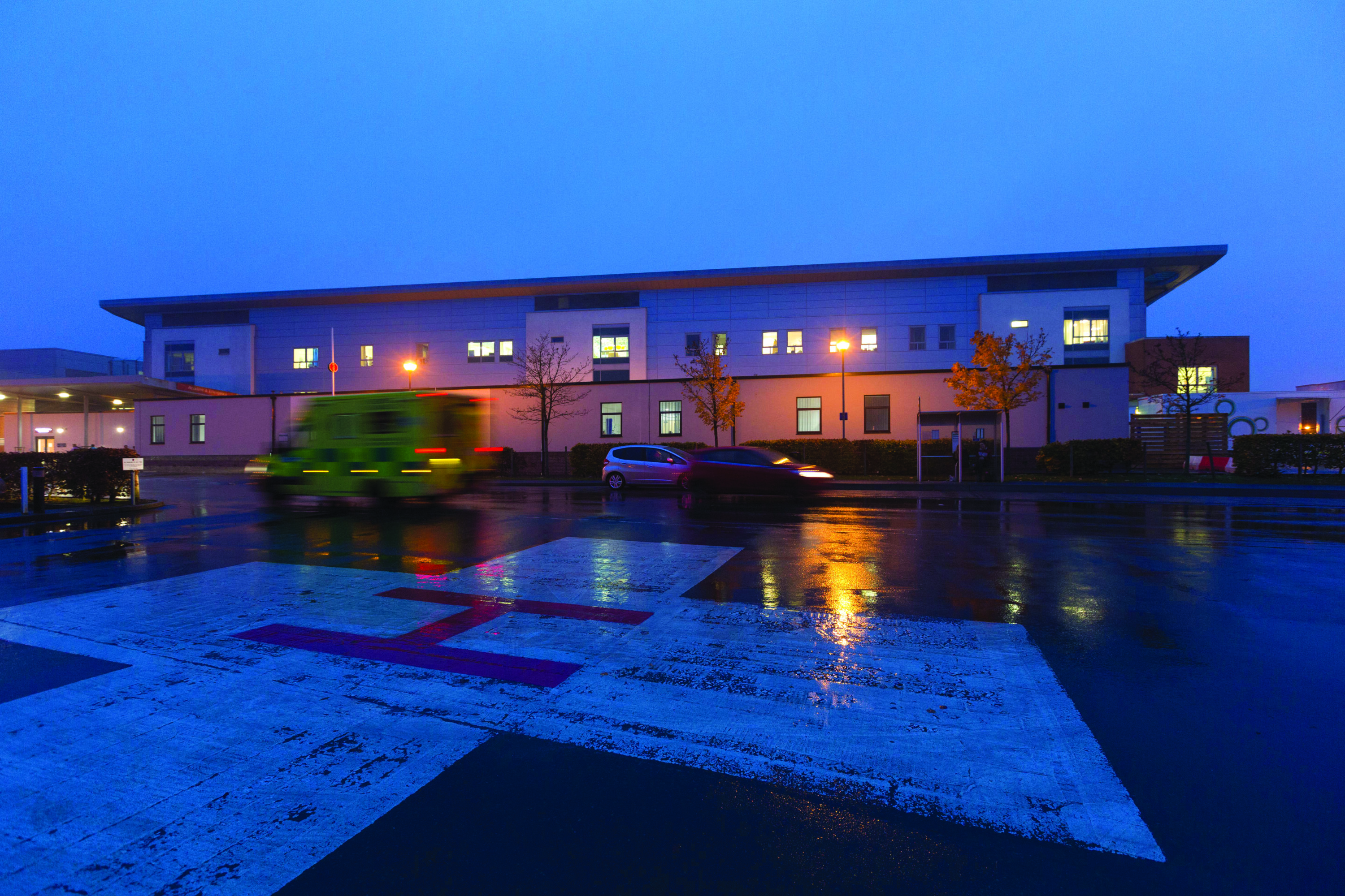 A picture of the Acute Medical Unit from outside. It is night time and the lights are on. There are cars in front of the building. In the foreground is a helicopter landing pad.