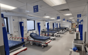 The inside of the Ambulance Receiving Centre. The room is predominantly white. Bed bays line both sides, but only the seven bays on the left hand side are clearly visible. Each bay has a blue mattress on a hospital bed, a blue curtain to act as a divider and a frosted window for light.