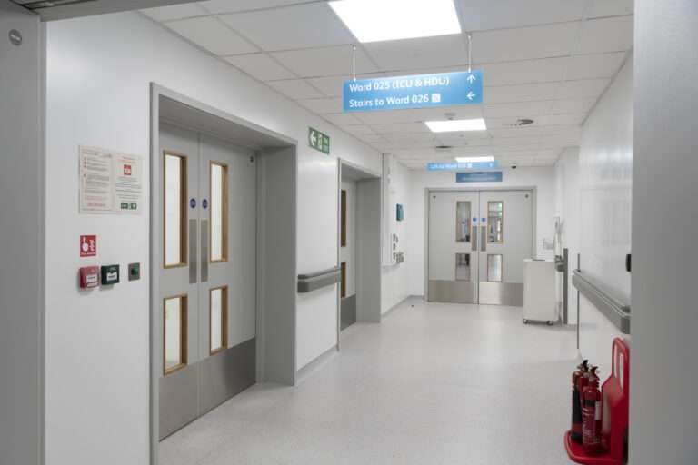 A corridor outside a ward. Everything is finished in white or very light grey. It is well lit. There are double doors to the left hand side and to the end of the corridor.