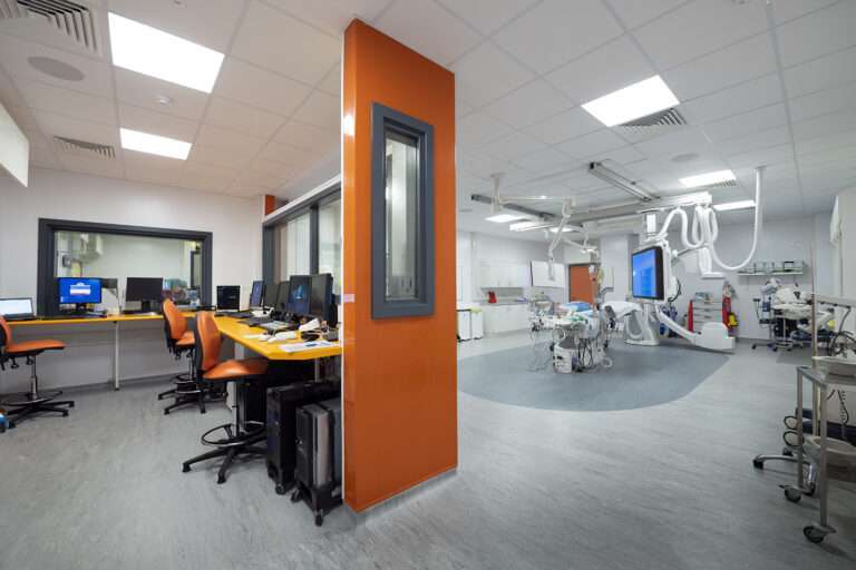 A cardiac catheterisation lab at Basingstoke. The walls and ceiling are white, the floor is a mottled grey vinyl. To the left there is a staff station with an orange dividing wall that features large glass windows. To the right there is the procedure area, which contains a bed for the patient as well as a variety of medical and diagnostic equipment.