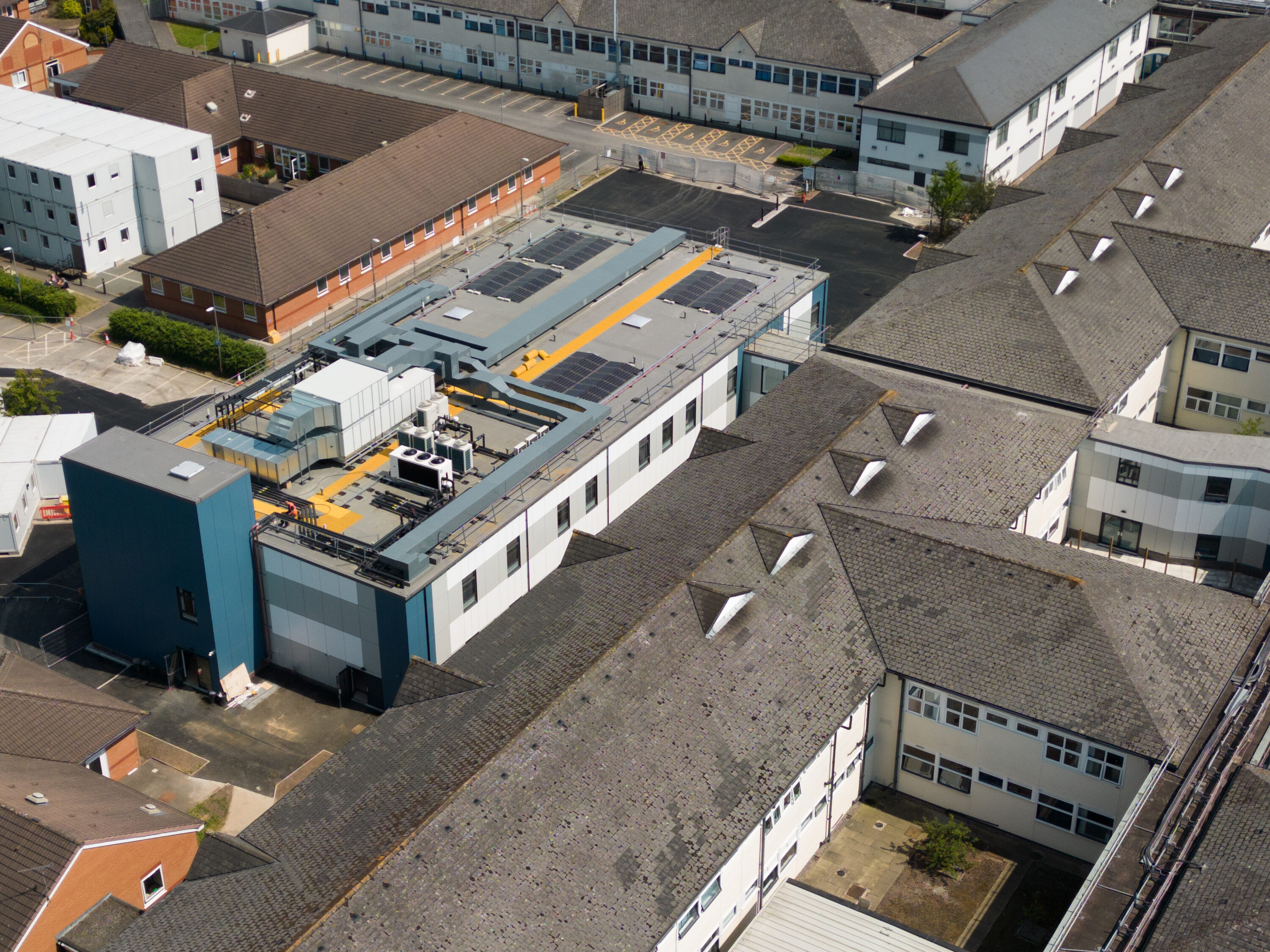 A three-quarters overhead view of the new ward building at Leighton Hospital, showing a range of plant machinery on the roof. This includes four photovoltaic solar panel arrays and a number of HVAC and air-conditioning units. Surrounding the new ward building are the older brick buildings of the hospital estate. The new ward building is finished in blue, grey and white cladding.