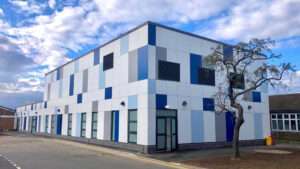 The exterior of the operating theatre building at Grantham. The entire building is clad with a mix of blue, grey and white cladding panels, while the lower floor features regularly spaced window and glass doors, all of which are frosted. To the right there is a large tree, and the sky is blue with white clouds.