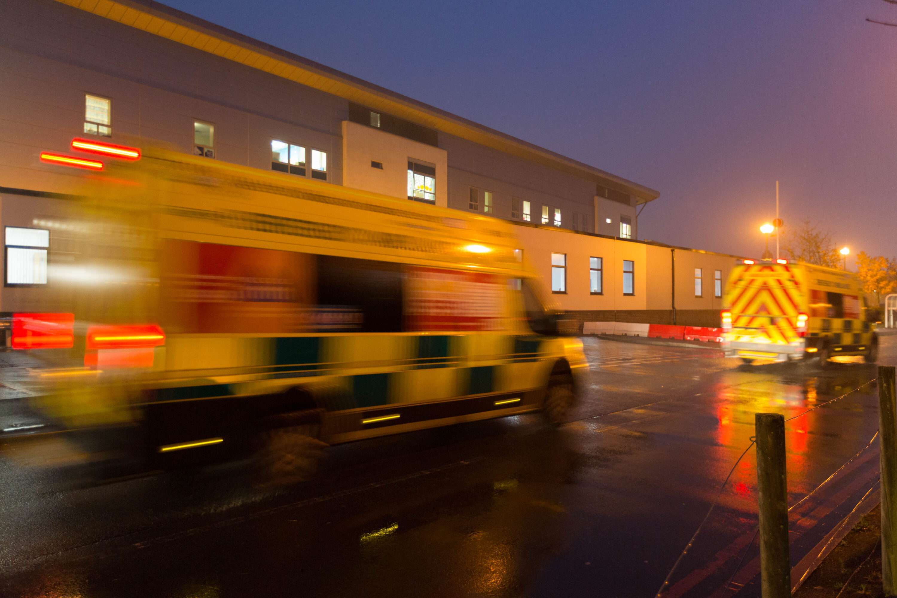 Two ambulances driving past a hospital ward at night time.