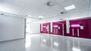 A newly finished, empty hospital ward. The floor is a very light grey vinyl, the walls and ceiling are white. To the left there is a large window and handwash station, to the right there is a ruby red wall with three bed bays.