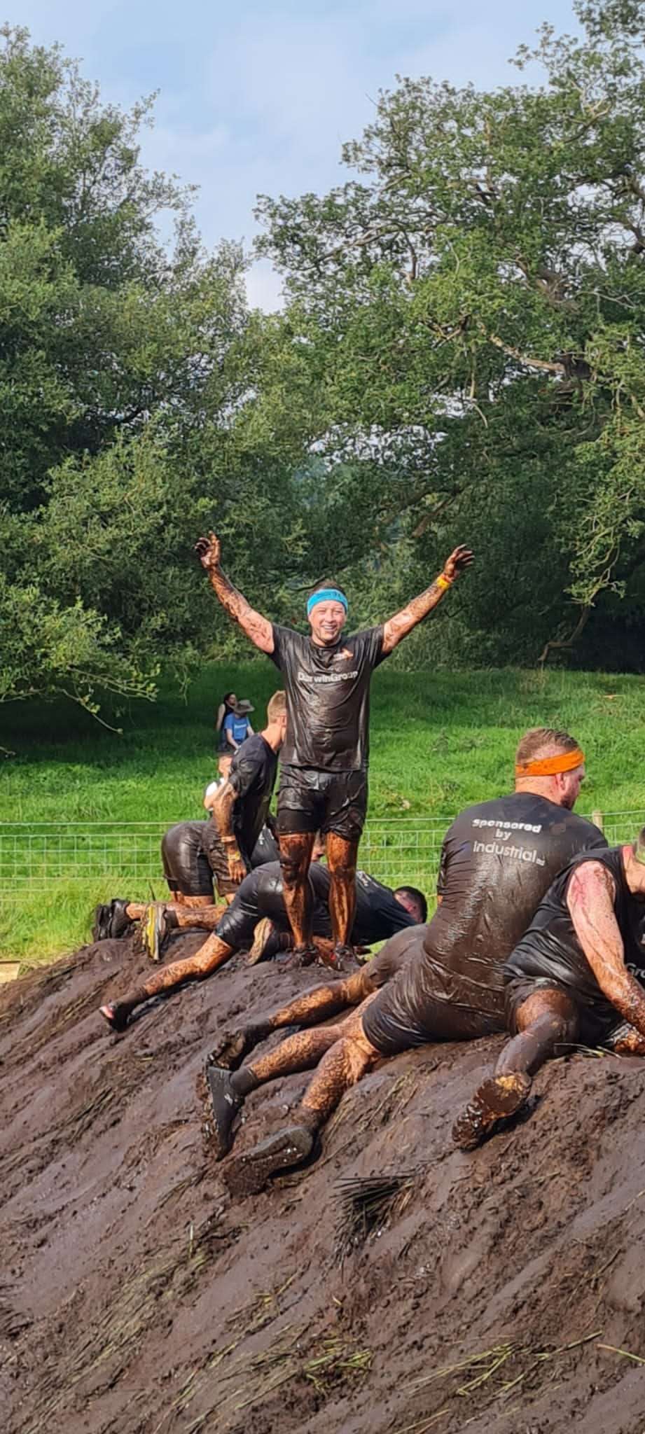A group of people tackling a very muddy obstacle as part of the Tough Mudder challenge. They are all soaking wet and covered in mud as they scramble over a mud bank. A Darwin Group employee is stood atop the mud bank with his arms raised in victory.