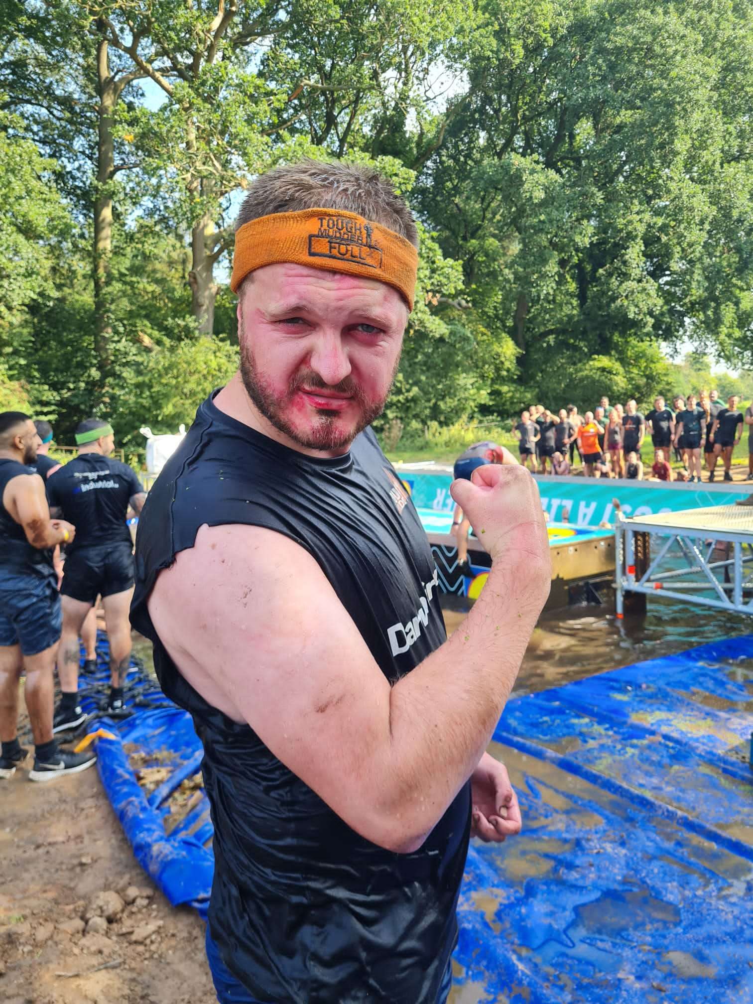 A Darwin Group employee in the middle of doing the Tough Mudder challenge. He is posing for the camera. In the background there is part of the obstacle course and a line of trees.