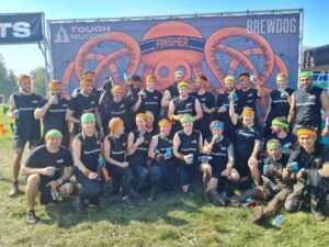 The Darwin Group team, having just finished the Tough Mudder challenge. There is a large banner behind them, the sky is blue and they are stood and sat on a grassy field.