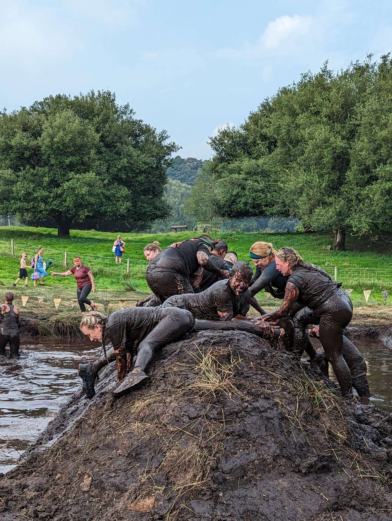 A group of Darwin Group employees tackling a very muddy obstacle as part of the Tough Mudder challenge. They are all soaking wet and covered in mud as they scramble over a mud bank with pools of water either side.