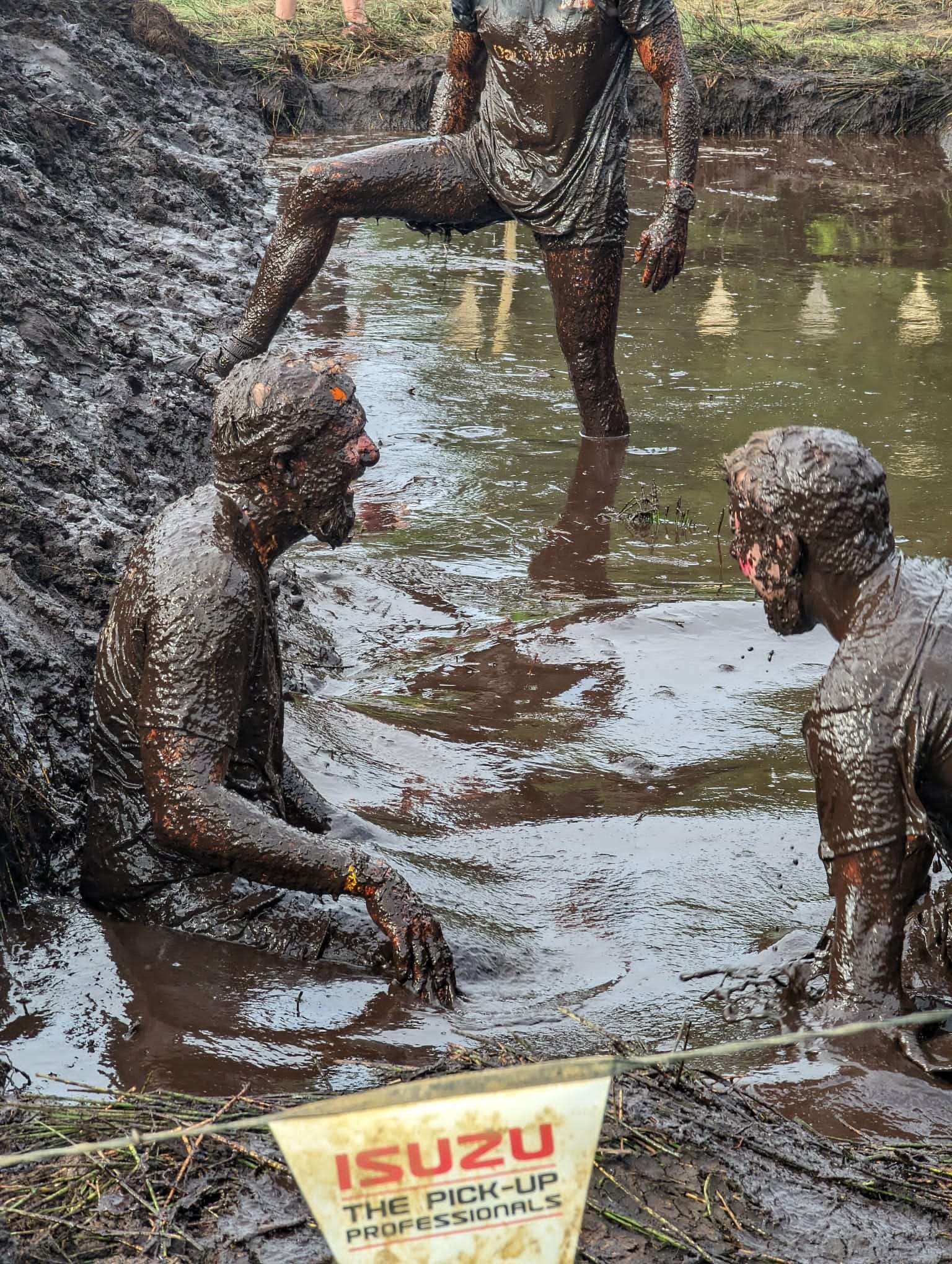 Three extremely muddy people stood in the middle of an obstacle consisting of a large mud bank and deep pools of water. The people are all covered entirely head to foot in mud.