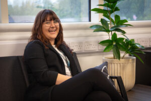 Photograph of a young woman sat down, looking at the camera and smiling. To the right is a large potted plant, there is a large window behind her.