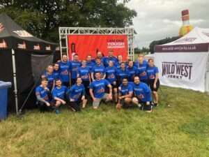 The Darwin Group team, ready to start the Tough Mudder challenge. There is a large banner behind them, gazebos to either side, the sky is overcast and they are stood in a grassy field.