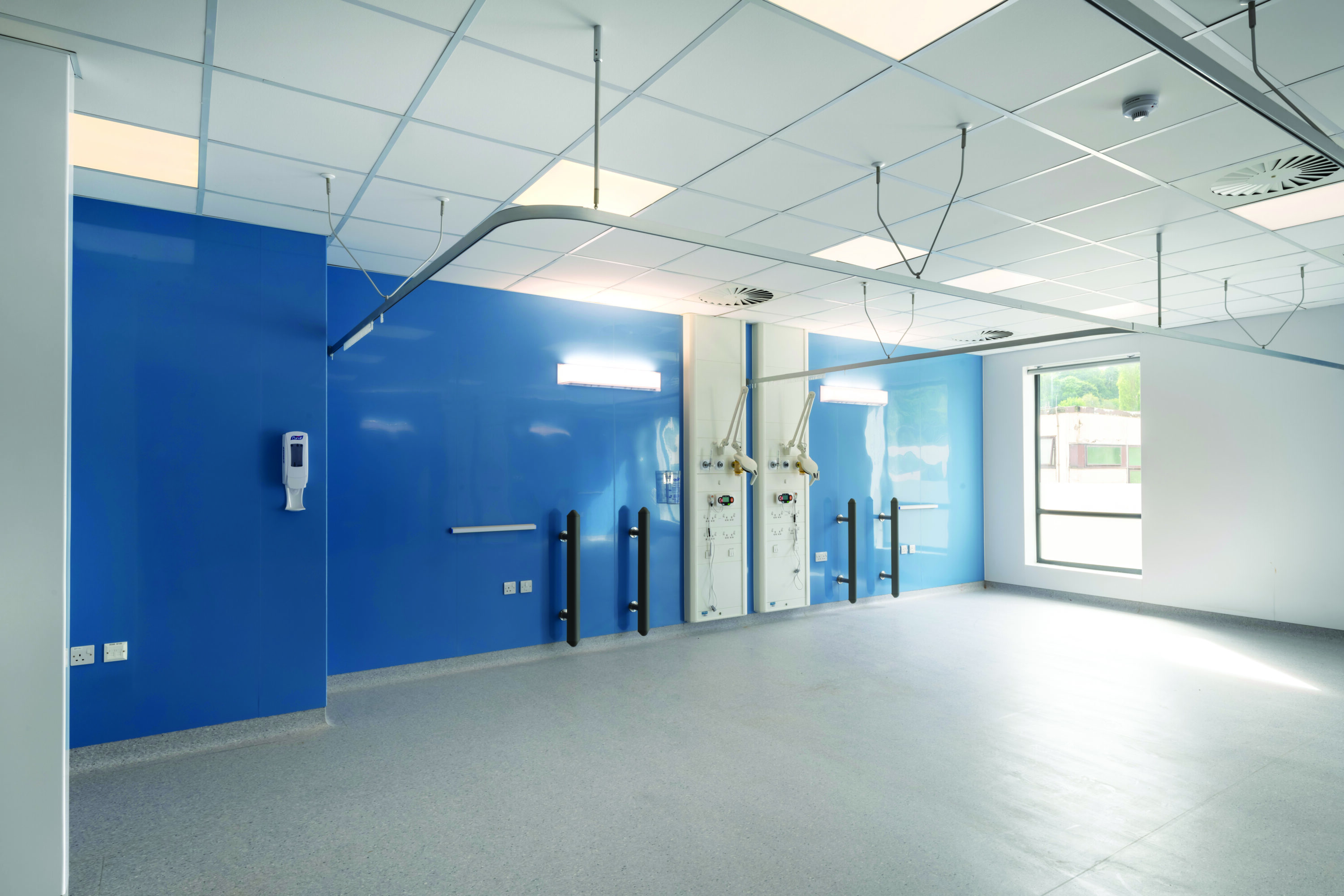 A newly finished, empty and well lit ward room. The rearmost wall is blue while the others are white, there is a large window to the right and the floor is a grey mottled vinyl.