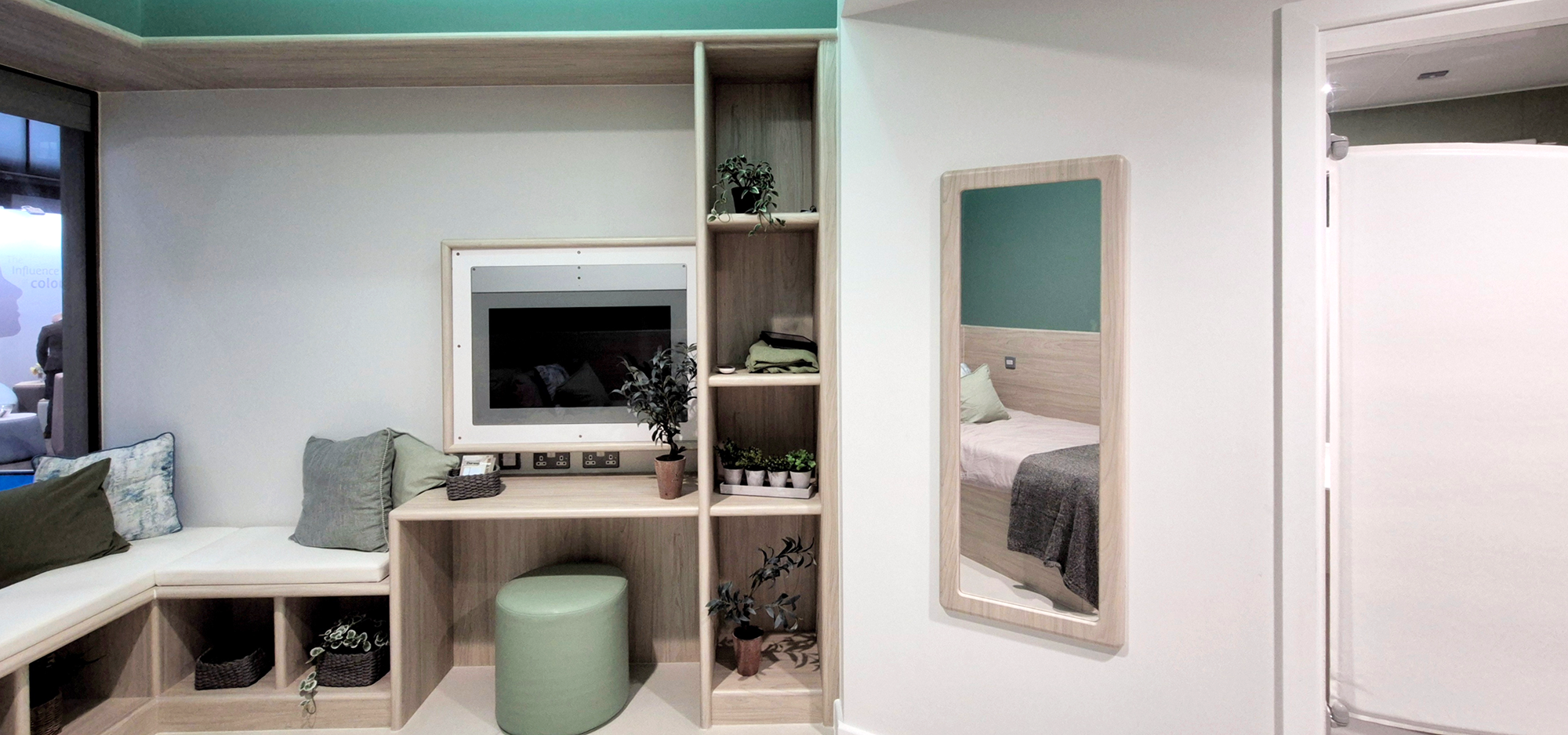 A wide angle photograph of the bedroom, showing the far wall with its integrated digital screen, shelving, desk and window seat